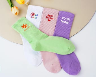 May include: A collection of colorful socks, each with unique text. The socks are in shades of green, white, pink, and purple. Text includes "Bride 8.17.25", "Best Mama", and "Your Name". A flower and logo are also visible.
