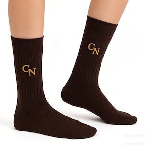 K&ouml;nnte beinhalten: Ein Paar dunkelbraune gerippte Socken mit den gestickten goldenen Buchstaben CN auf der Au&szlig;enseite jeder Socke. Die Socken haben eine vertikale Rippenstruktur und eine durchgehend dunkelbraune Farbe mit verst&auml;rktem Zehen- und Fersenbereich.