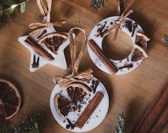 May include: Three white, scented ornaments in star, circle, and round shapes, decorated with dried orange slices, cinnamon sticks, and cloves. Each has a brown ribbon for hanging, set against a wooden background with greenery.