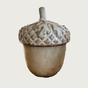 May include: A ceramic acorn-shaped container. The cap is light brown and textured with a diamond pattern, while the body is off-white and smooth. It has a small stem. This decorative item is likely used for storage or display.