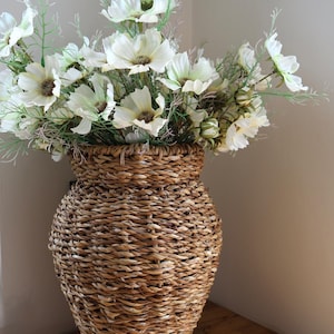 Peut inclure: Un grand vase en jonc de mer tressé marron avec un corps large et arrondi et un col étroit, rempli d'un grand arrangement de fleurs de cosmos artificielles blanches aux centres sombres et aux tiges et feuillages vert clair.