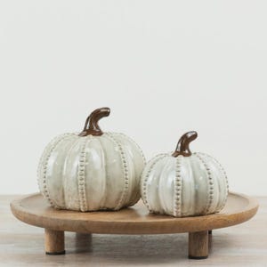 May include: Two ceramic pumpkins with a glossy off-white finish and raised beaded detailing along the ribs, each topped with a smooth, dark brown stem, displayed together on a round, light-toned wooden pedestal stand with three short legs.