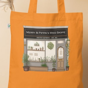 Może przedstawiać: Pomarańczowa torba na zakupy z czarno-białą ilustracją witryny sklepowej z tekstem "Merry & Pippin's Weed Shoppe" i "South Farthing -  KSE 301".