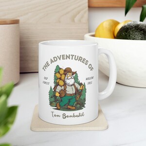 May include: White ceramic mug with a cartoon illustration of a man carrying a bundle of pine cones. The text "The Adventures of Tom Bombadil" is printed on the mug.