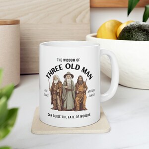 Könnte beinhalten: Eine weiße Keramiktasse mit einer schwarzen und braunen Illustration von drei alten Männern mit langen weißen Bärten. Der Text "THE WISDOM OF THREE OLD MAN" ist in Schwarz um die Illustration herum gedruckt. Der Text "THE SHIRE" ist unter dem linken Mann gedruckt, "MIDDLE EARTH" unter dem rechten Mann und "CAN GUIDE THE FATE OF WORLDS" unter der Illustration.
