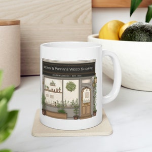 Könnte beinhalten: Weiße Keramiktasse mit einer Schwarzweißillustration einer Ladenfront. Der Laden heißt "Merry & Pippin's Weed Shoppe" und befindet sich in "South Farthing, Est. 2021".