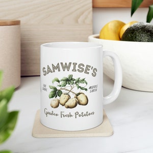May include: White ceramic mug with a brown graphic of potatoes and a green plant. The text "Samwise's" is at the top of the mug, with "The Shire" and "Middle Earth" on either side of the plant. The text "Garden Fresh Potatoes" is at the bottom of the mug.