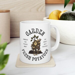 Könnte beinhalten: Eine weiße Keramiktasse mit einer Schwarzweißillustration einer Kartoffelfigur, die Kartoffeln hält. Der Text "Garden Fresh Potatoes" ist in einem schwarzen Kreis um die Illustration gedruckt. Der Text "Samwise "The Potato Man" und "The Shire's Best Potatoes" ist in schwarzer Schrift auf der Tasse gedruckt.
