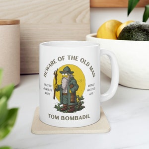 May include: White ceramic mug with a cartoon illustration of a bearded man with a hat and a staff. The text reads "Beware of the Old Man" and "Tom Bombadil".