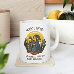 Könnte beinhalten: Weiße Keramiktasse mit einem gelben Cartoon-Bild von drei Figuren aus Herr der Ringe. Der Text "Where's Frodo?" befindet sich oben auf der Tasse. Der Text "Sam Gamgee, Pippin and Merry Brandybuck" befindet sich unten auf der Tasse.