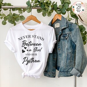 Pode incluir: Camiseta branca com texto preto que diz "Never Stand Between a Girl and Her Python". O texto está em uma fonte cursiva e inclui três pequenos corações.