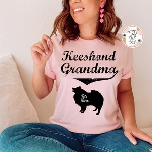 Può includere: Maglietta rosa con il testo "Keeshond Grandma" e una silhouette nera di un cane Keeshond con il testo "Nome dell'animale" sotto.
