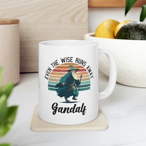 Könnte beinhalten: Weiße Keramiktasse mit einer Retro-Grafik von Gandalf aus Der Herr der Ringe, der davonläuft. Der Text lautet "Selbst die Weisen laufen weg Gandalf".