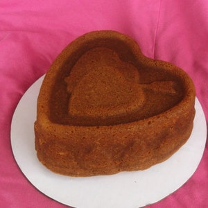 May include: A heart-shaped cake with a brown, slightly textured surface. The cake is sitting on a white cake stand.