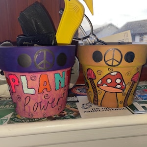 Puede incluir: Dos macetas de terracota pintadas, una morada con las palabras "Plant Power" y la otra amarilla con un signo de paz y hongos.