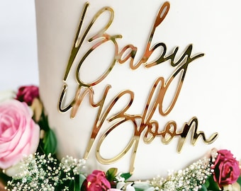 May include: Gold glitter cake topper with the words "Baby in Bloom" in a cursive script font.