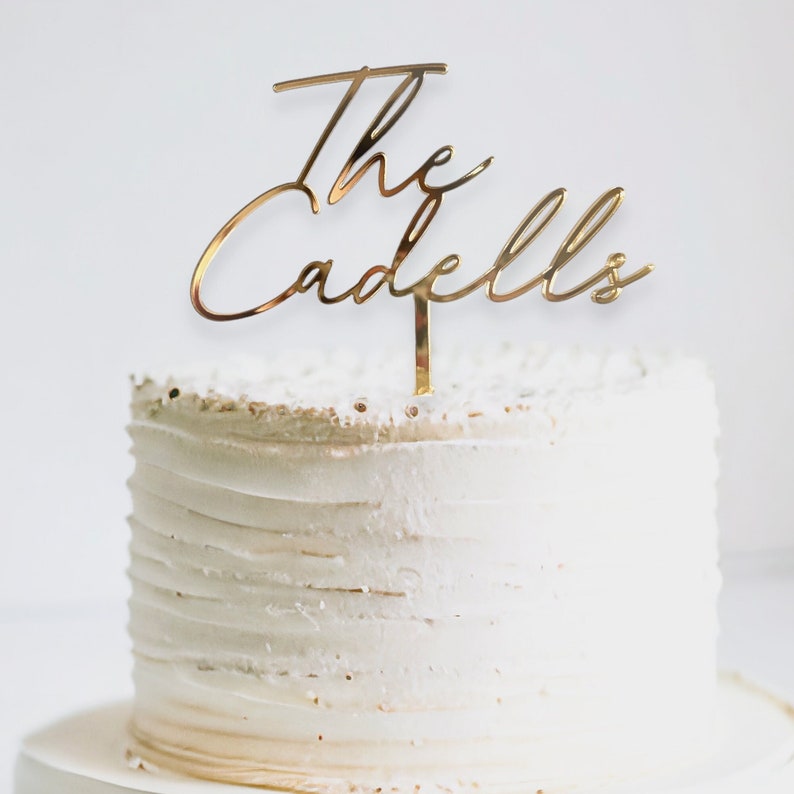 May include: A white cake with gold lettering on top that reads "The Cadells".