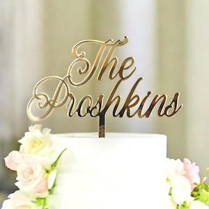 May include: A gold-colored cake topper with the words "The Proshkins" in an elegant script. The topper is placed on a white cake decorated with white and pink flowers, perfect for a wedding or anniversary celebration.