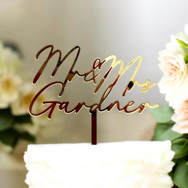 May include: Gold mirror acrylic cake topper with the text "Mr & Mrs Gardner" in a cursive script font.