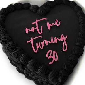 May include: A heart-shaped black cake with black frosting and decorative swirls around the edge. The cake has pink text that reads "not me turning 30".