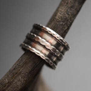 May include: A wide silver band ring featuring a dark, oxidised finish and three horizontal rows of textured, scalloped detailing. The metal surface displays a weathered appearance with high-contrast highlights along the raised edges.