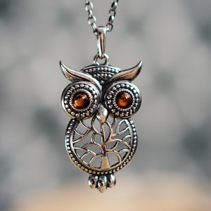 Può includere: Una collana con ciondolo a forma di gufo color argento con due pietre rotonde color ambra come occhi. Il corpo del gufo presenta un dettagliato design ad albero in filigrana sul petto, sospeso a una catena in metallo abbinata a maglie.