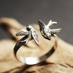 Op de afbeelding: Een zilverkleurige ring met twee gedetailleerde zwaluwontwerpen. De vogels zijn aan weerszijden van de ring geplaatst, met uitgespreide vleugels. De ring staat tegen een vage achtergrond, waardoor de details van het sieraad worden benadrukt.