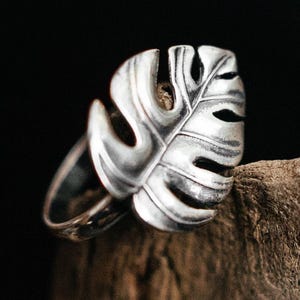 Könnte beinhalten: Ein silberner Ring mit einem detaillierten Monstera-Blatt-Design. Das Blatt hat komplizierte Ausschnitte, vor einem dunklen Hintergrund. Die Textur und Form des Rings werden hervorgehoben. Der Ring wird auf einem Holzstück präsentiert.