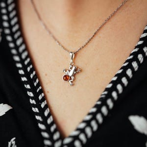 以下が含まれることがあります： 琥珀の石が付いたカエルのペンダントがついたシルバーネックレス。カエルは後ろ足で座り、前足は伸ばされています。