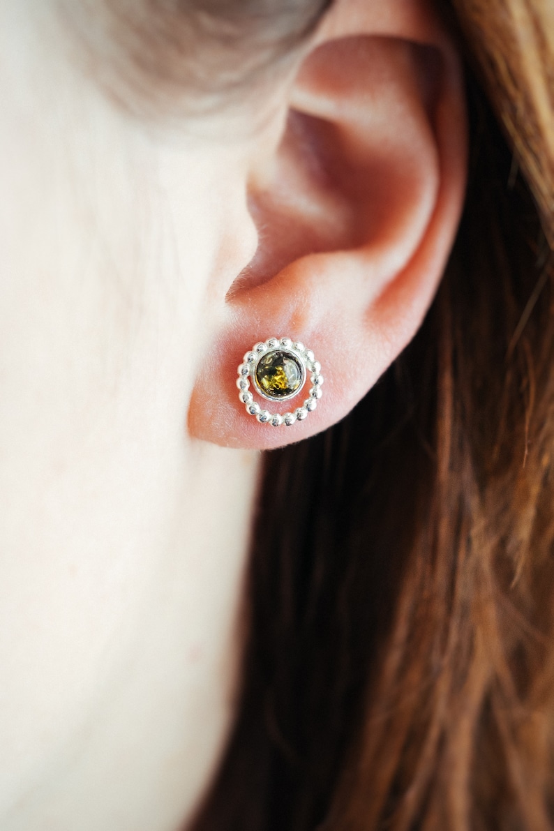 May include: A pair of silver earrings with a small green gemstone in the centre of each earring. The earrings are in the shape of a circle with a beaded border.
