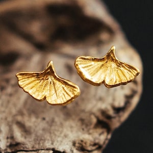 Op de afbeelding: Een paar goudkleurige oorstekers in de vorm van ginkgo bladeren. De oorbellen hebben gedetailleerde aderen en een gepolijste afwerking, die het licht reflecteert. De oorbellen zijn geplaatst tegen een vage, neutrale achtergrond.