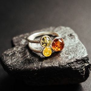 Può includere: Un anello d'argento con tre pietre di ambra. Le pietre sono di diverse tonalità di giallo e arancio. L'anello è su una roccia grigio scuro.