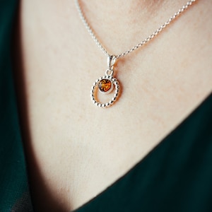 May include: A silver necklace with a round pendant featuring a small amber stone in the centre. The pendant is surrounded by a circle of small silver beads.