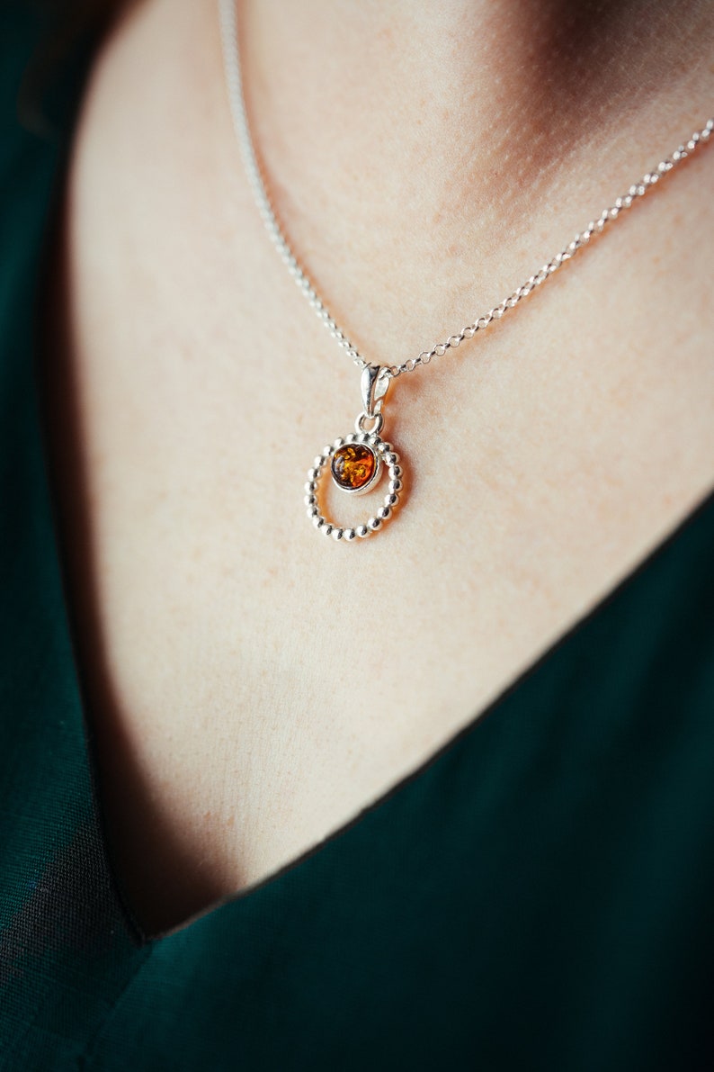 May include: A silver necklace with a round pendant featuring a small amber stone in the centre. The pendant is surrounded by a circle of small silver beads.