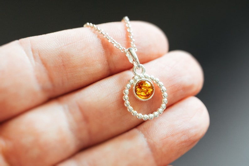 May include: A silver necklace with a round pendant featuring a small amber stone. The pendant is surrounded by a circle of small silver beads.