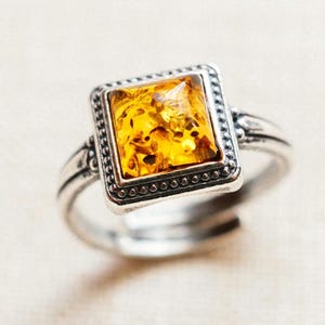 Könnte beinhalten: Ein silberner Ring mit einem quadratischen Bernstein. Der Bernstein hat eine warme gelbe Farbe mit sichtbaren Einschlüssen. Der Ring hat eine detaillierte, verzierte Fassung mit einem Perlenrand. Schmuck.