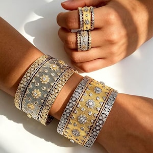 May include: Two wide cuff bracelets and two stacked rings featuring a mixed metal design of silver and gold tones. The jewelry pieces are decorated with intricate filigree patterns, textured surfaces, and small embedded clear stones.