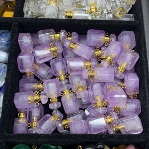 May include: A selection of small, amethyst-coloured perfume bottles with gold-coloured caps. The rectangular bottles are arranged in a black tray, with other gemstones visible in the background. The caps have a small loop for attaching a chain or charm.