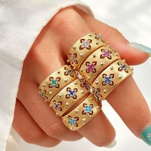 May include: A set of wide gold-toned rings with a textured, brushed finish. Each ring features a repeating pattern of four-petaled flower motifs, with each petal inlaid with small, colorful gemstones in shades of blue, pink, green, and purple.