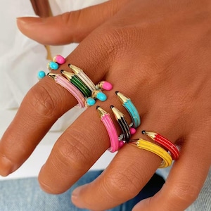 Pode incluir: Uma coleção de anéis em forma de lápis em miniatura, com cores vibrantes de esmalte, incluindo rosa, verde, branco, azul, preto, amarelo e vermelho. Cada anel tem uma ponta dourada e uma pequena conta colorida na extremidade do lápis.