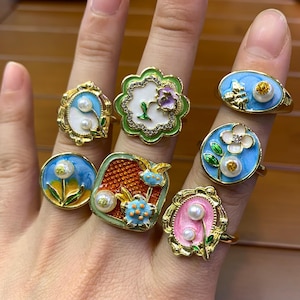 May include: A collection of gold-toned rings with colorful enamel designs. Each ring features unique floral or abstract motifs, including pearls, flowers, and geometric shapes. The rings showcase a variety of colors, such as blue, green, pink, and white, with intricate detailing.