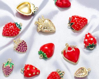 Puede incluir: Una colección de dijes en forma de fresa en varios diseños. Algunos son de color rojo liso, otros dorados con pedrería o gemas rojas. Hay detalles verdes y dorados en las hojas y los tallos. Estos dijes son probablemente para hacer joyas.
