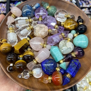 May include: A selection of gemstone perfume bottles in a variety of shapes and colours, including rose quartz, amethyst, and lapis lazuli. Each bottle has a gold-coloured cap and is displayed in a wooden bowl. The gemstones are polished and reflect light.