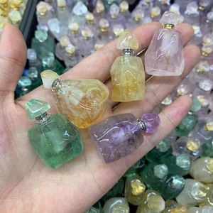 May include: A selection of gemstone perfume bottles in shades of green, yellow, purple, and pink. Each bottle features a silver-coloured metal top. The bottles are held in a hand, with many more similar bottles visible in the background.
