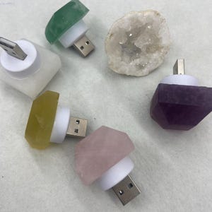 Puede incluir: Una colección de cargadores USB con tapas de piedras preciosas en varios colores, incluyendo verde, rosa, morado y amarillo. Los cargadores tienen una base blanca y un conector USB. También hay una geoda.
