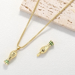 Könnte beinhalten: Eine goldfarbene Halskette mit einem Anhänger in Form eines Pokals, der an der Basis und am oberen Rand mit grünen Emaille-Streifen versehen ist. Der Anhänger hängt an einer strukturierten Goldkette, daneben liegt ein passender Pokal-Anhänger.
