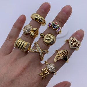 Könnte beinhalten: Eine Sammlung goldfarbener Ringe, jeder mit einem einzigartigen Design. Zu den Stilen gehören ein Band mit eingebetteten Steinen, ein herzförmiger Ring mit filigranem Design und ein Ring mit einer Zeichentrickfigur. Einige Ringe haben klare Steine.