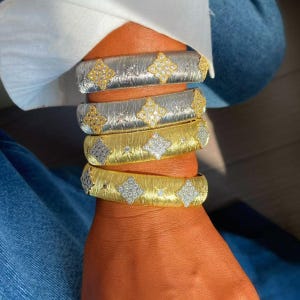 Op de afbeelding: Een stapel van vier brede armbanden met een textuur van geborsteld metaal. Twee armbanden zijn zilverkleurig met goudkleurige ruitvormige accenten, en twee zijn goudkleurig met zilverkleurige accenten, allemaal bezet met kleine heldere steentjes.