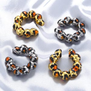 Puede incluir: Cuatro puños para la oreja con diseño de estampado de leopardo. Dos son dorados y dos plateados. Cada puño está compuesto por pequeñas cuentas redondas con manchas negras, naranjas y marrones. Los puños están dispuestos sobre una superficie de satén plateado.