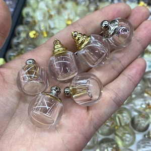 May include: A selection of miniature, transparent glass perfume bottles with gold and silver-toned lids. The bottles are spherical with a rectangular base, held in a hand. The lids feature decorative detailing.